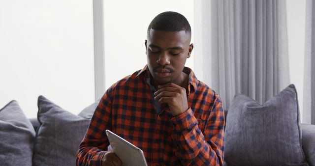 A young African American man in a plaid shirt is sitting on a couch using a digital tablet with a thoughtful expression. This image can be used for technology ads, articles about relaxation or home life, and content related to digital tools and modern lifestyles.