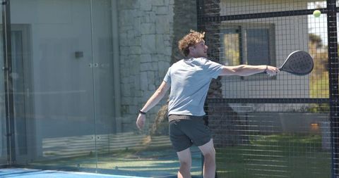 Padel player swinging paddle and reaching for yellow ball on sunny glass court