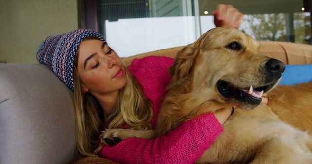 A woman in a knit cap and sweater is lying on a couch with her arms around a golden retriever dog. Both appear relaxed and content. This image can be used in articles and advertisements related to pet care, companionship, lifestyle, and home comfort.
