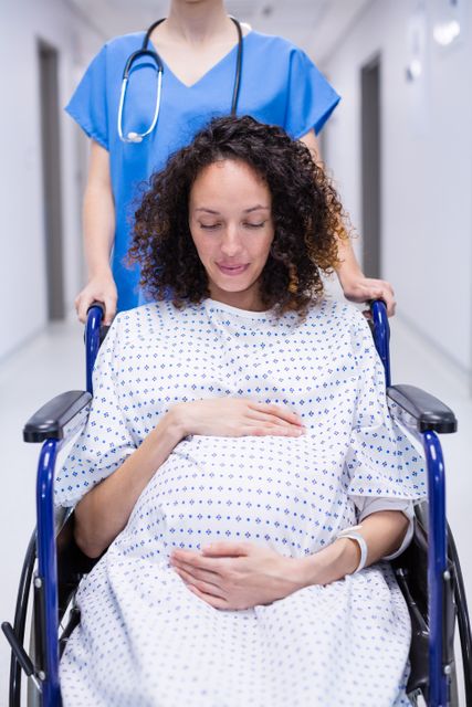 Pregnant woman sitting in wheelchair, gently touching her belly, being pushed by nurse in hospital corridor. Ideal for use in healthcare, maternity, pregnancy, and medical care contexts. Can be used in articles, brochures, and websites related to prenatal care, hospital services, and patient care.
