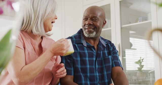 Senior couple is smiling and spending quality time together in a bright kitchen. Both appear happy and engaged in conversation. This image is perfect for use in articles or advertisements related to senior living, relationships in later life, and healthy lifestyles for older adults.