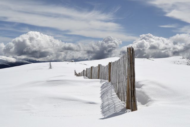 Picture of a serene winter landscape featuring a rustic wooden fence stretching across the snow-covered ground under a sky filled with dramatic clouds. Ideal for use in articles or advertisements promoting outdoor activities, winter vacations, or environmental conservation. Useful for backgrounds, greeting cards, and calendars showcasing natural beauty and tranquility.