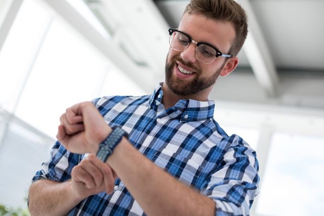 Young male executive in a modern office environment using a smartwatch. He is wearing casual business attire, including a blue plaid shirt and glasses. This image can be used for business technology, productivity, and modern workplace themes.