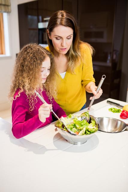 Mother and daughter are preparing a fresh salad together in a modern kitchen. This image can be used for promoting family bonding activities, healthy eating habits, parenting tips, and home cooking recipes. It is ideal for websites, blogs, and advertisements focused on family life, nutrition, and culinary arts.
