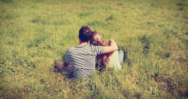 A young Caucasian couple is sitting closely together in a grassy field, embracing and enjoying a moment of intimacy, with copy space. Their relaxed posture and the serene outdoor setting suggest a romantic and peaceful connection.