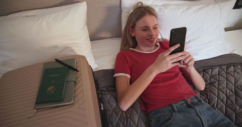 Teen Girl Using Smartphone with Suitcase and Passport Nearby in Bedroom