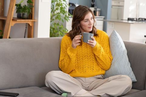 Cozy at-home moment woman in mustard sweater relaxing on sofa with mug and phone