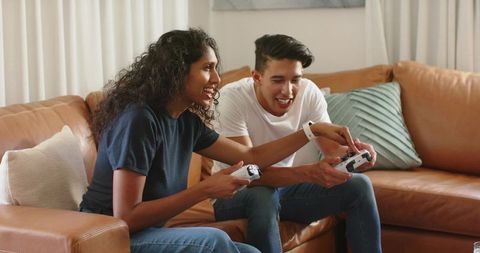 African american couple playing video games on leather sofa, smiling and laughing