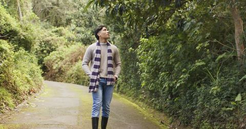 Asian man walking mossy forest lane wearing striped scarf, knit sweater and boots
