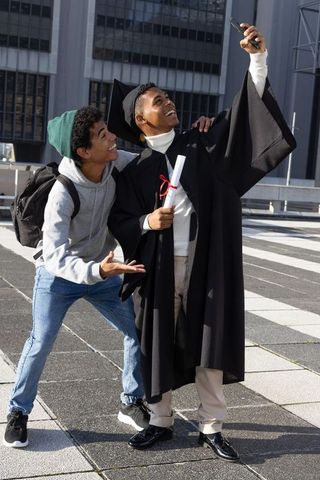 African american graduate celebrating with friend taking selfie on urban campus