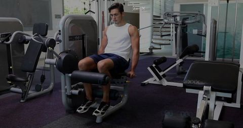 Fit man performing leg extension on machine in modern gym with spiral staircase
