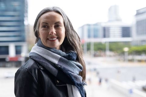 Mature woman smiling in urban plaza wearing leather jacket and blue-gray scarf looking right