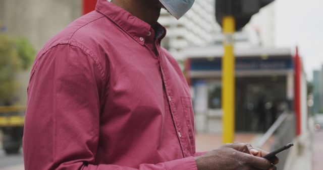 Man wearing face mask and red shirt using smartphone outdoors in city. Useful for content on urban life, communication, public health safety, and technology use in public spaces.