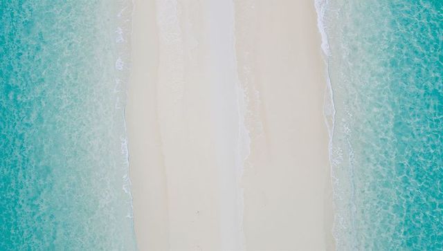 Aerial central sandbar splitting turquoise seas with white foam and faint footprints