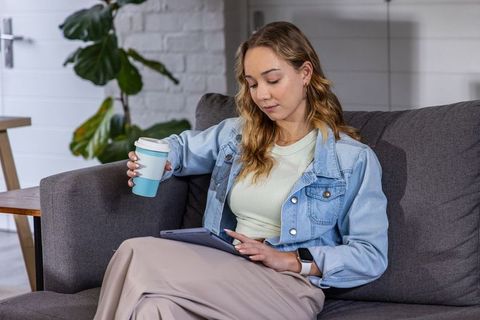 Young woman relaxing on sofa holding reusable cup and using tablet for remote work home lifestyle