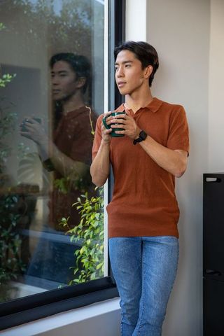 Contemplative asian man standing by window holding mug, modern minimal interior, daylight greenery