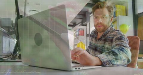 Bearded man typing on laptop in small office with yellow hard hat and analytics overlay