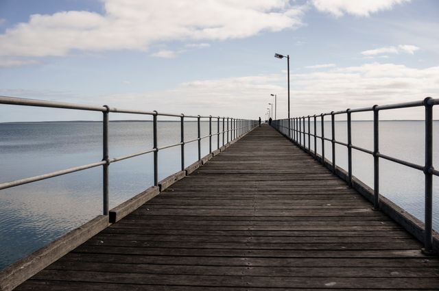 Long wooden pier stretches into a calm blue sea under a partly cloudy sky. Ideal for representing tranquil coastal landscapes, travel destinations, serene ocean views, and outdoor relaxation. Useful for blogs, travel guides, vacation advertisements, and coastal lifestyle themes.