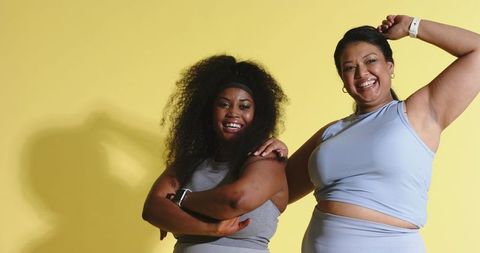 Two women smiling and posing in activewear wearing smartwatches on bright yellow backdrop