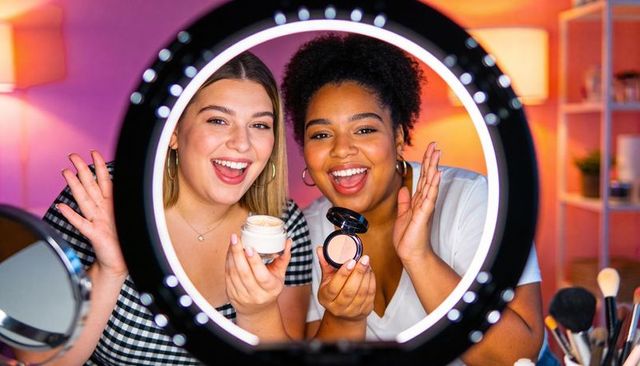 Two beauty creators filming makeup tutorial with ring light holding cream and compact powder