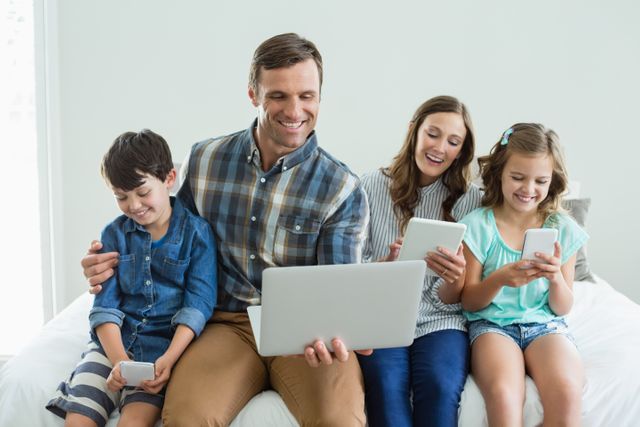 Smiling family using laptop, digital tablet and mobile phone in bedroom at home