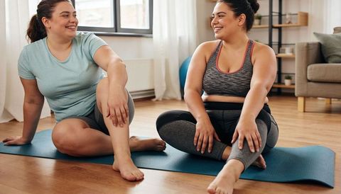 Two women stretching on blue mats at home smiling chatting and playfully nudging