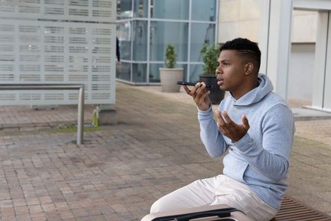 African-american traveler recording voice note on phone while sitting with luggage