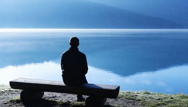 Man sitting on bench facing calm reflective lake at dawn for solitude and reflection