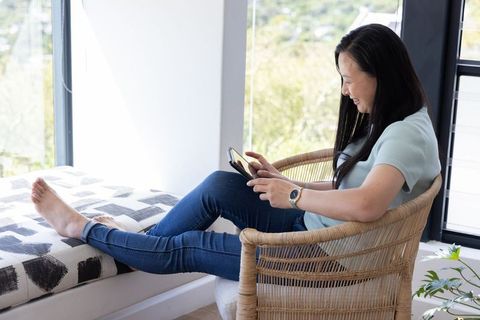 Asian mature woman relaxing at home using smartphone by sunlit window for digital life