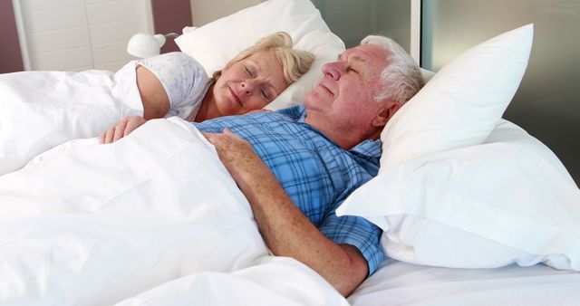 A senior couple peacefully sleeping together in a cozy bed. The scene portrays comfort, rest, and togetherness with the soft white blankets and pillows adding a sense of cozy warmth. This image is ideal for use in articles or advertisements related to senior living, wellness, healthy sleep habits, relationships, or home bedding products.
