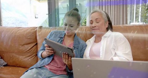 Denim teen sharing tablet as senior leaning over laptop on sofa, multigenerational bonding
