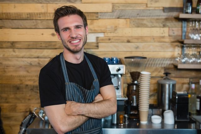 Young male waiter standing confidently with arms crossed in a cozy café. Ideal for use in articles or advertisements related to hospitality, customer service, coffee shops, and professional work environments.