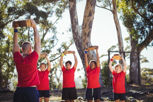 Trainer leading group of kids in obstacle course training, each child holding a wooden log overhead. Ideal for use in articles or advertisements about children's fitness, teamwork activities, outdoor exercises, summer camps, and physical education programs.