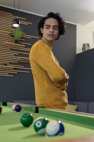 Young man leaning on pool table wearing mustard sweater studying billiard shot