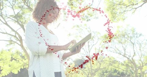 Smiling woman tapping tablet in sunlit park with floating heart overlay and lens flare