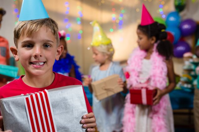 Boy holding a wrapped gift with friends in background at a birthday party. Children wearing party hats and colorful decorations create a festive atmosphere. Ideal for use in content related to children's parties, celebrations, joy, and childhood memories.