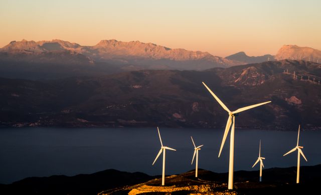 Wind turbines standing on a picturesque mountainous hillside bathed in the light of the setting sun. Ideal for topics related to renewable energy, environmental conservation, and sustainable development. Suitable for articles, presentations, and marketing materials about clean energy solutions, eco-friendly practices, and beautiful natural landscapes.