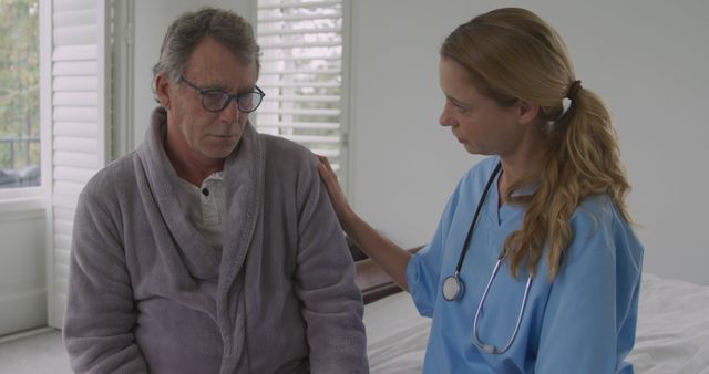 Caregiver wearing scrubs and stethoscope comforting elderly man in robe beside bed in bedroom. Perfect for healthcare, elderly care support, home nurse services, compassionate care, and medical assistance themes.