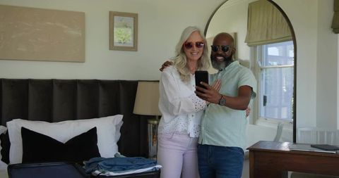 Senior couple taking selfie in cozy bedroom with open suitcase, sunglasses and travel vibe