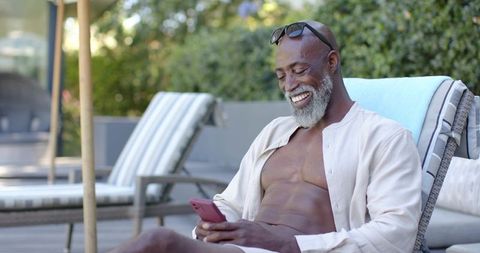 Smiling african american senior relaxing poolside using smartphone, sunglasses, open shirt