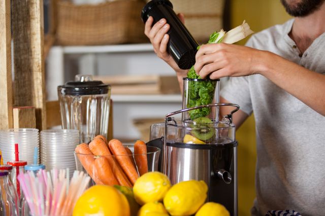 Male staff member preparing fresh juice using a juicer at a health grocery shop. Fresh vegetables and fruits like carrots, lemons, and celery are visible. Ideal for use in content related to healthy living, organic food, nutrition, wellness, and grocery stores.