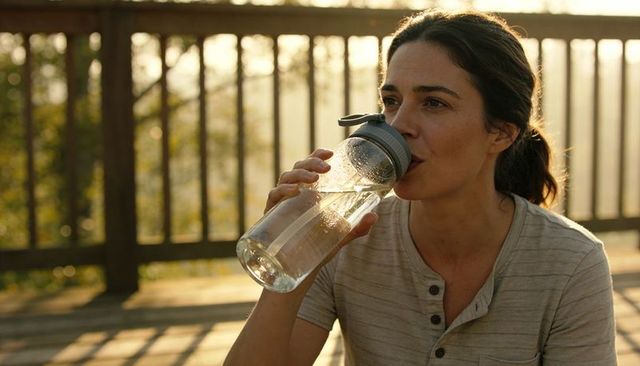 Woman drinking water from reusable bottle on sunlit wooden deck during golden hour