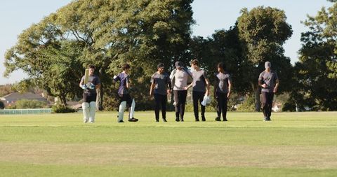 Diverse cricket team walking across pitch during batting practice with coach signalling
