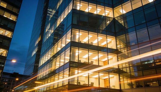 Glowing glass office tower showing lit workspaces at night with traffic light trails