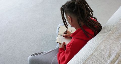 Man in red hoodie journaling on carpet next to sofa with dreadlocks minimal neutral room