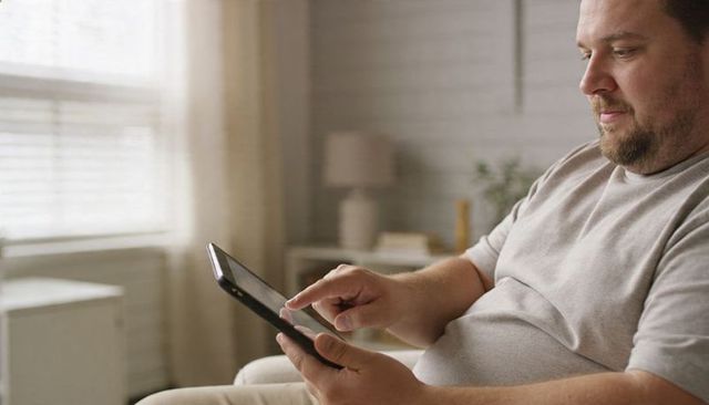 Casual man tapping tablet while relaxing on couch near sunlit window in cozy living room