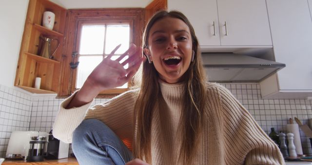 Young woman smiling and greeting, wearing a casual sweater in a modern, well-lit kitchen with wooden accents. Suitable for themes related to lifestyle, happiness, domesticity, greetings, and casual moments at home. Ideal for content about friendly communication, everyday scenes, home decor, and modern living.