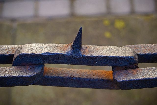 Close-up of a rusted old metal chain with a spike link, captured against a blurred pavement background. This gritty, industrial image is suitable for themes of durability, strength, age, and weathering. It can be used in articles about historical architecture, industrial photography, safety equipment, and as a background for rugged design elements.