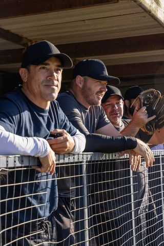 Baseball teammates leaning on dugout fence watching game holding glove and phone