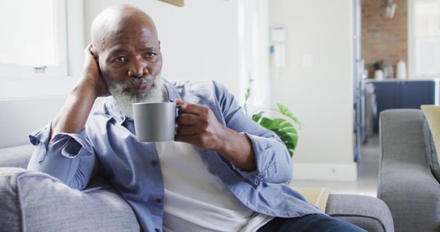 Senior man relaxing at home while drinking coffee. Sitting on sofa with cozy, peaceful expression. Use for lifestyle, retirement, relaxation, morning routines, and home comfort themes.